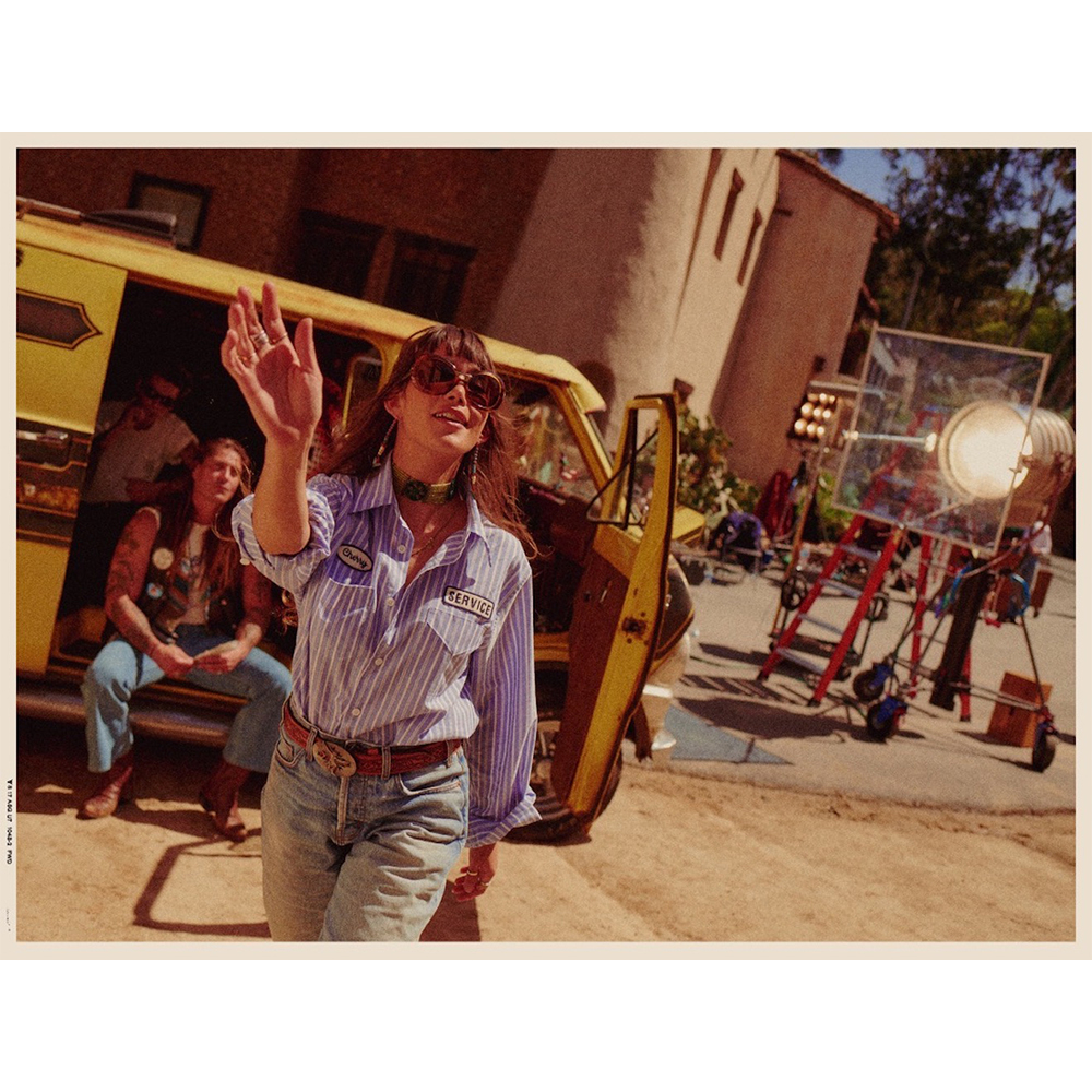 Woman waves in vintage-style outdoor shoot on film set with yellow van, film equipment, buildings, and other people visible in background.