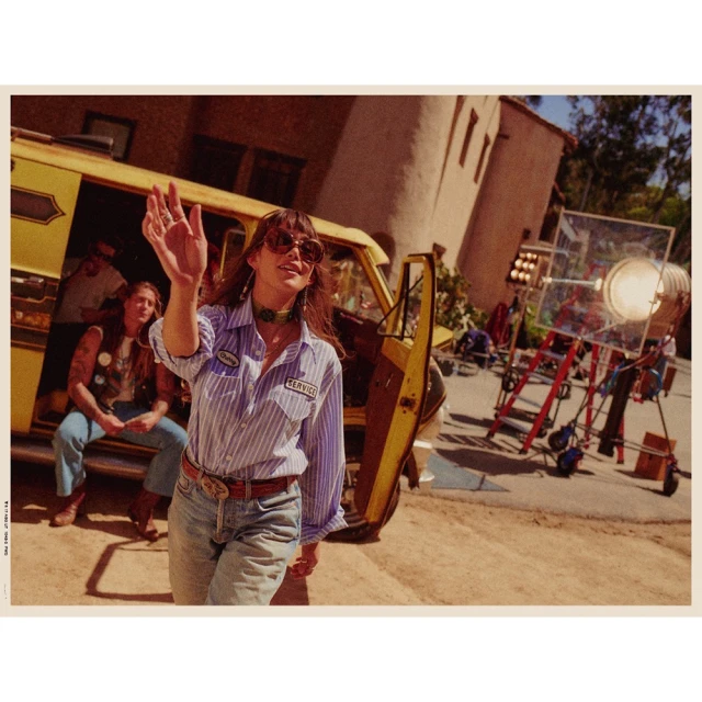 Woman waves in vintage-style outdoor shoot on film set with yellow van, film equipment, buildings, and other people visible in background.