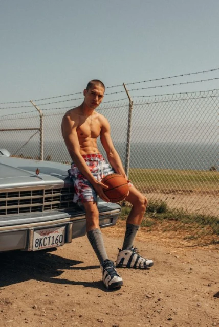 Athletic man with basketball sits on car hood. Shirtless model in colorful shorts, socks, and shoes poses outdoors in sunlight.