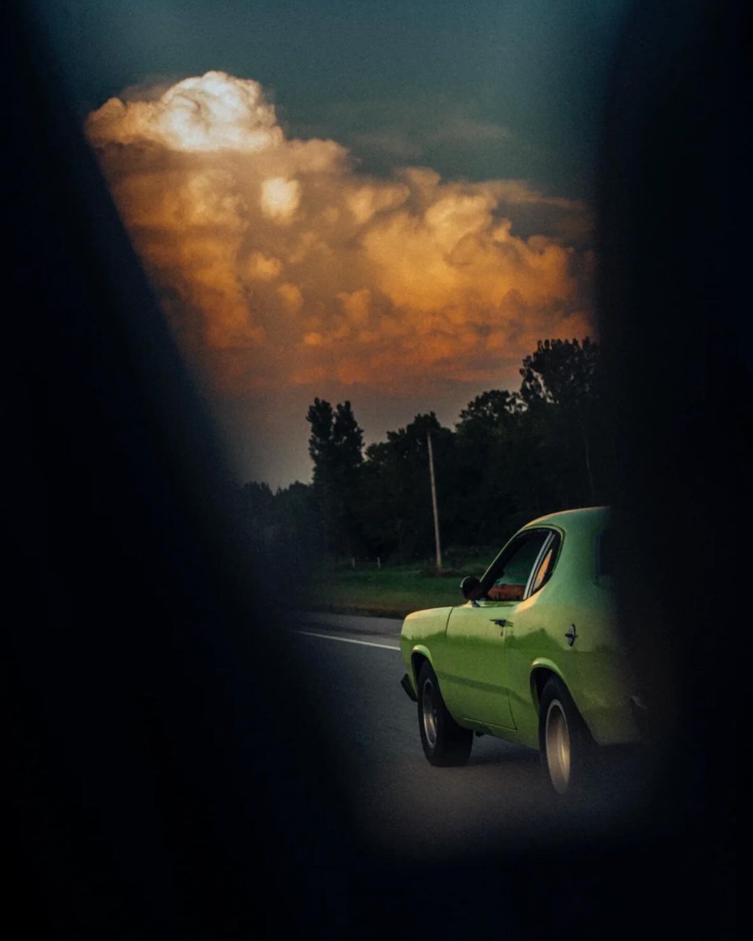 Vintage green car driving on a paved road under a dramatic sky with clouds. Scenic view of a classic automobile in motion.