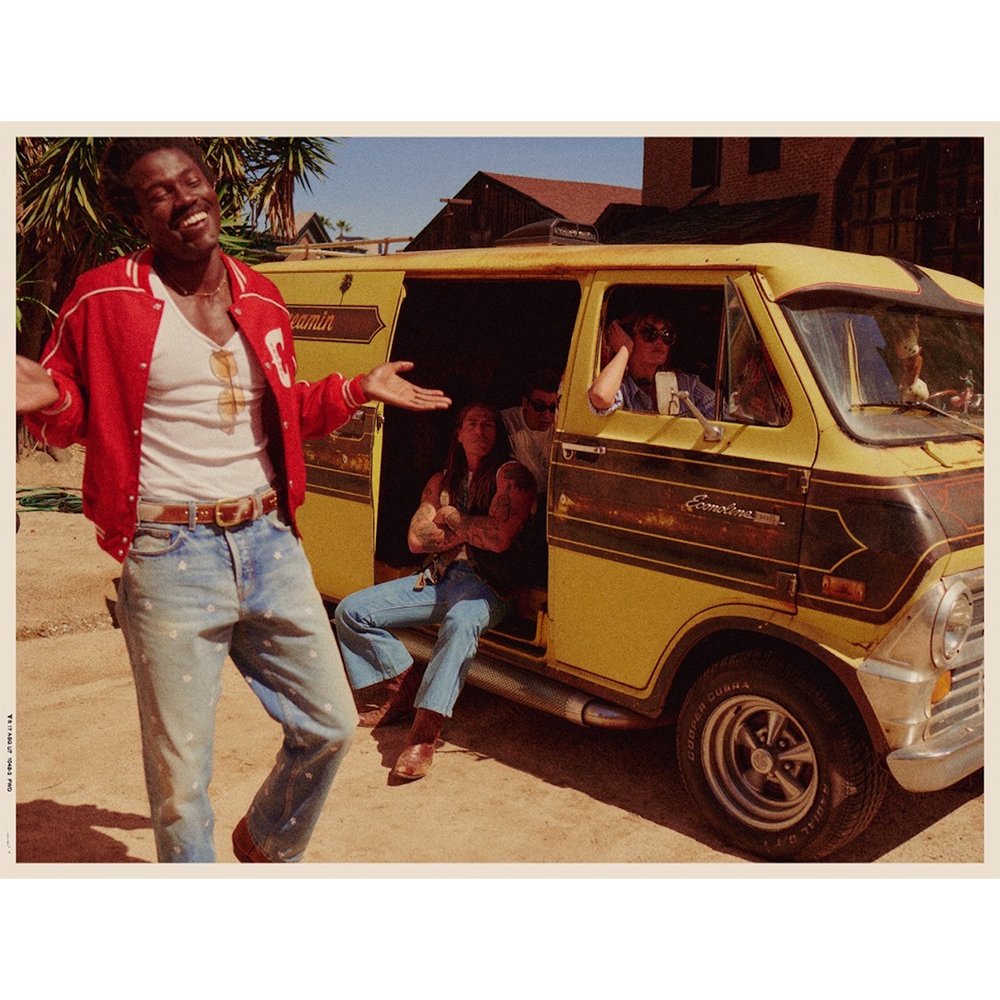 Group of people pose with a vintage yellow van outdoors. An African-American man in a red jacket stands smiling in the foreground.