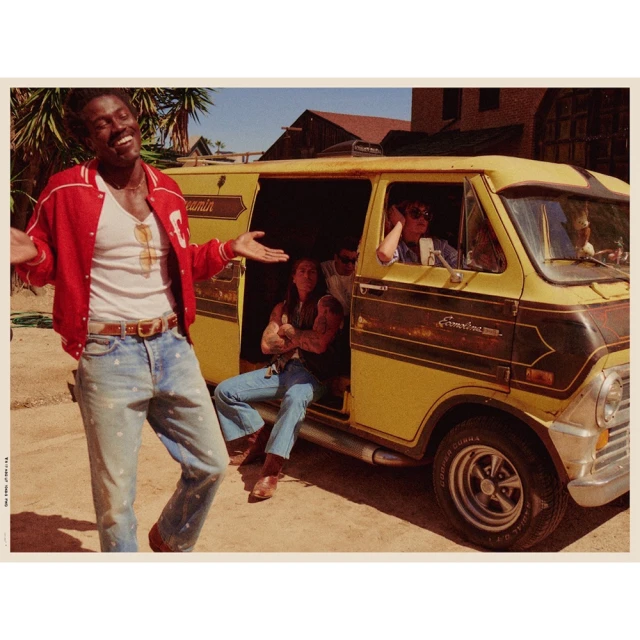Group of people pose with a vintage yellow van outdoors. An African-American man in a red jacket stands smiling in the foreground.