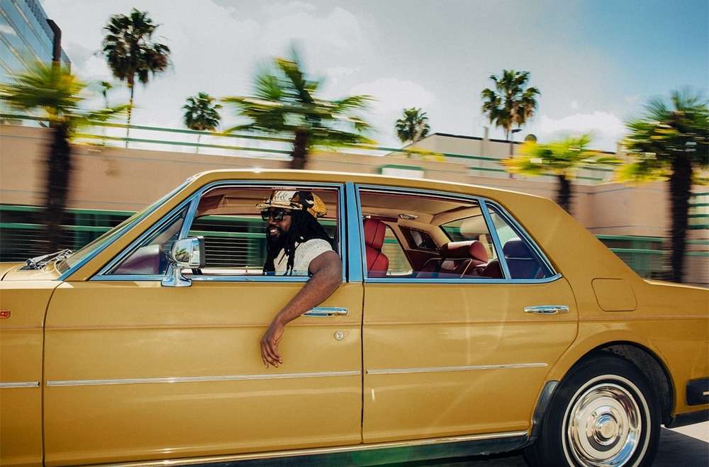 Man with dreadlocks drives vintage gold car along palm-lined street on a sunny day, wearing a hat and white shirt, arm out window.