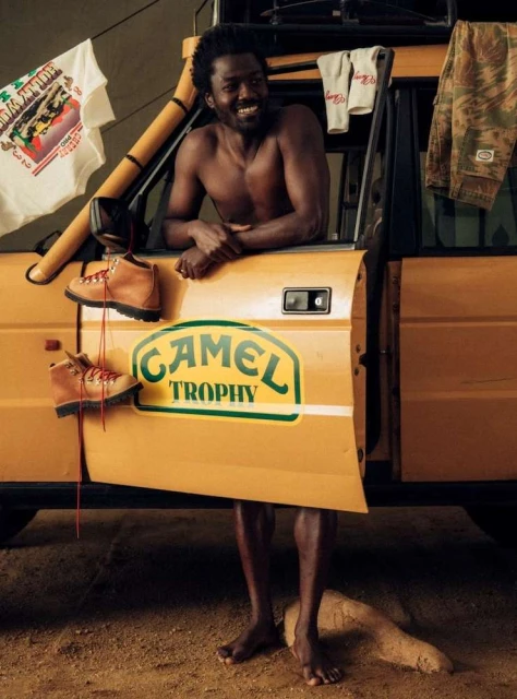 Shirtless African American man leans against yellow off-road vehicle with Camel Trophy logo, wearing shorts and smiling outdoors.