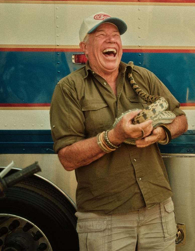 Elderly man laughing heartily while holding a patterned snake, wearing an olive shirt and baseball cap.