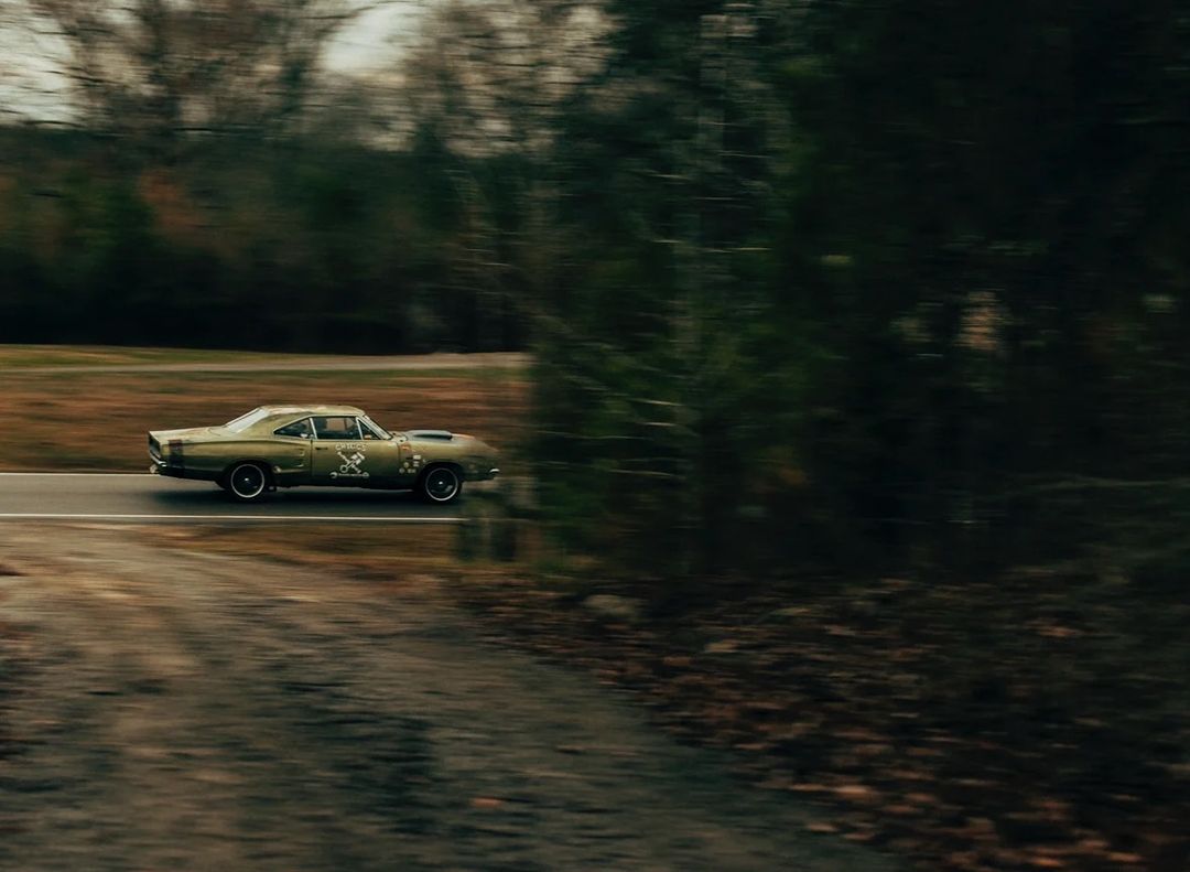 Vintage muscle car speeding down a rural road surrounded by trees, showcasing automotive design and natural scenery.