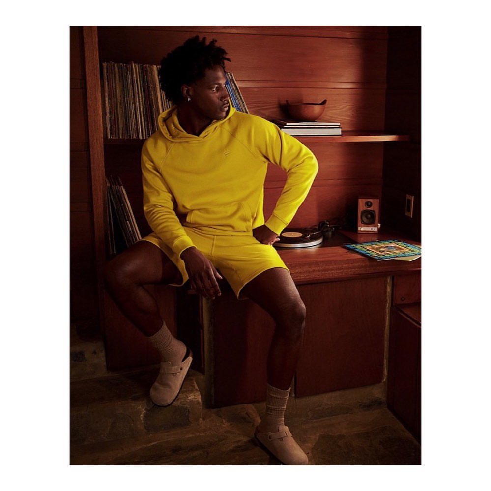 Young man in yellow hooded sweatshirt and shorts sitting on a wood desk in a wood-paneled room with vinyl records and a turntable.