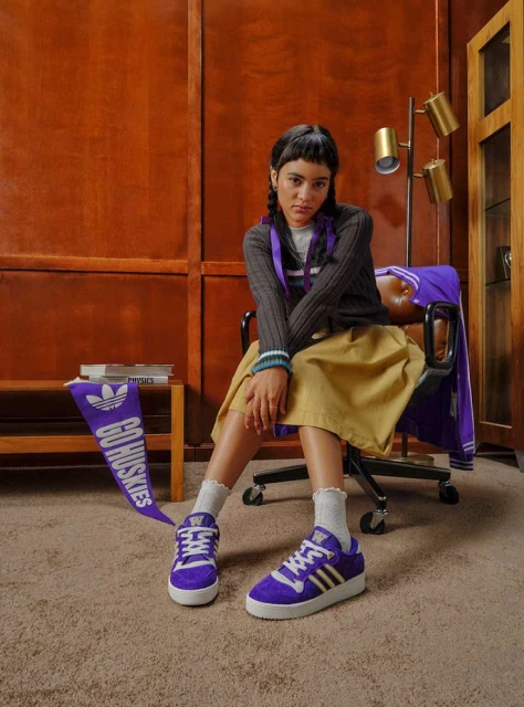 Young woman in casual clothing sits next to Adidas sneakers and a 'Go Huskies' banner in an indoor setting with carpet and wood panelling.