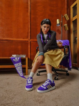 Young woman in casual clothing sits next to Adidas sneakers and a 'Go Huskies' banner in an indoor setting with carpet and wood panelling.