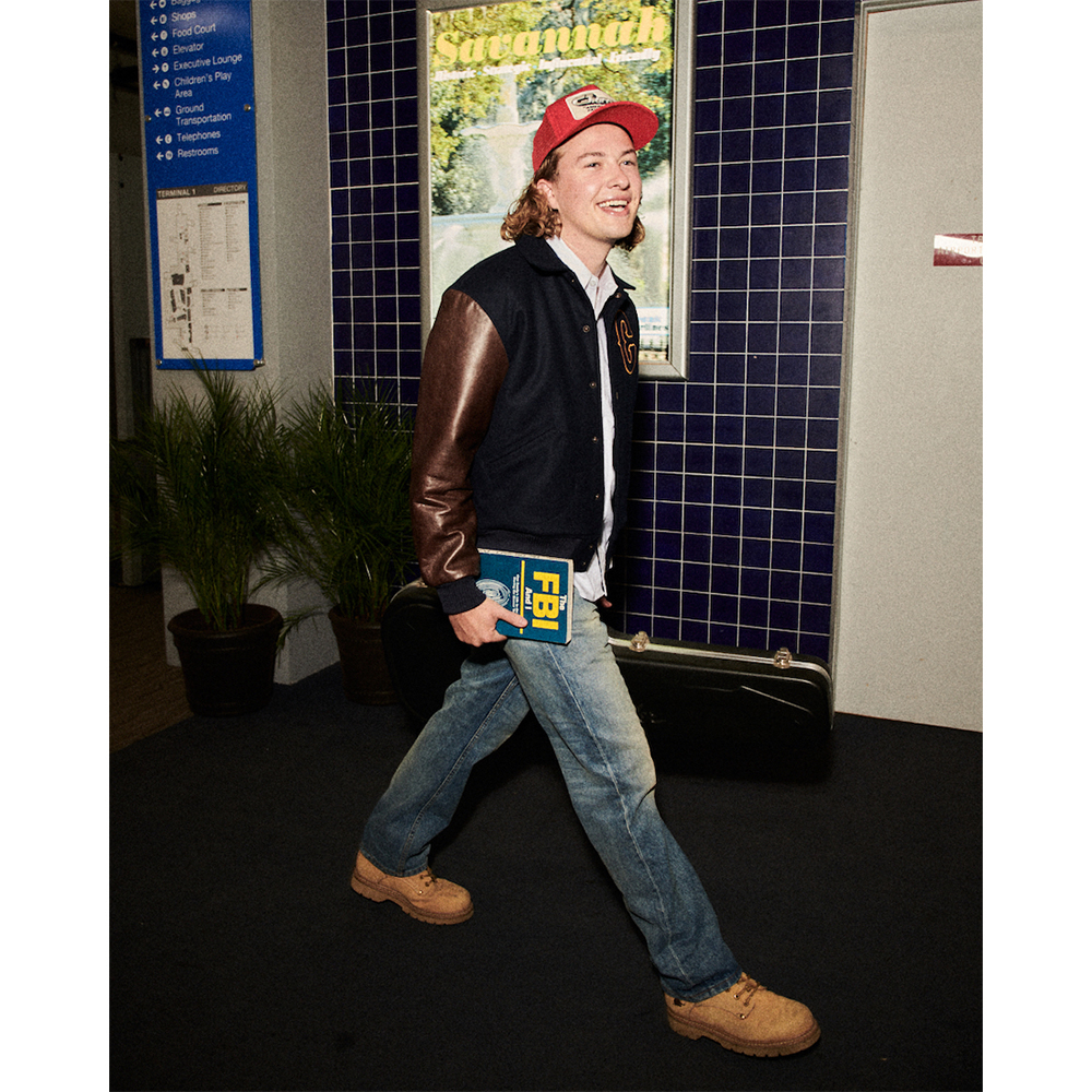 Young adult male walks in vintage style with a book, wearing a letterman jacket, jeans, red cap, and tan work boots, indoor environment.