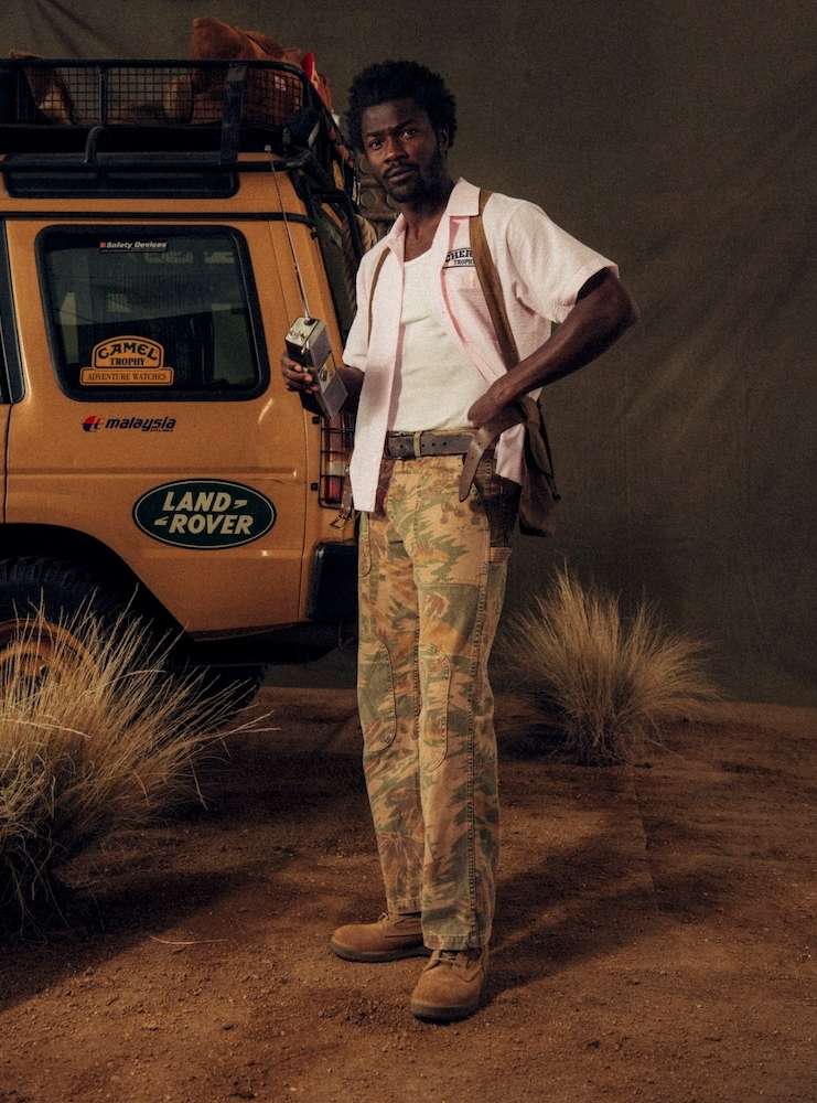 A man stands confidently near a Land Rover, holding a vintage radio, dressed in earth-toned pants, and a pink shirt, evoking an outdoor adventure.
