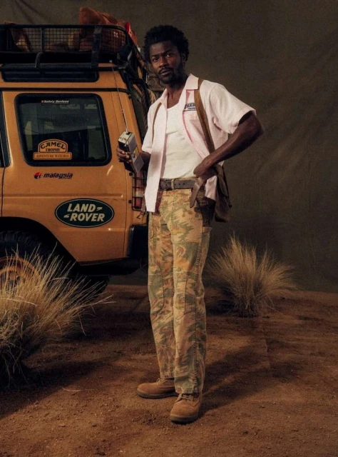 A man stands confidently near a Land Rover, holding a vintage radio, dressed in earth-toned pants, and a pink shirt, evoking an outdoor adventure.
