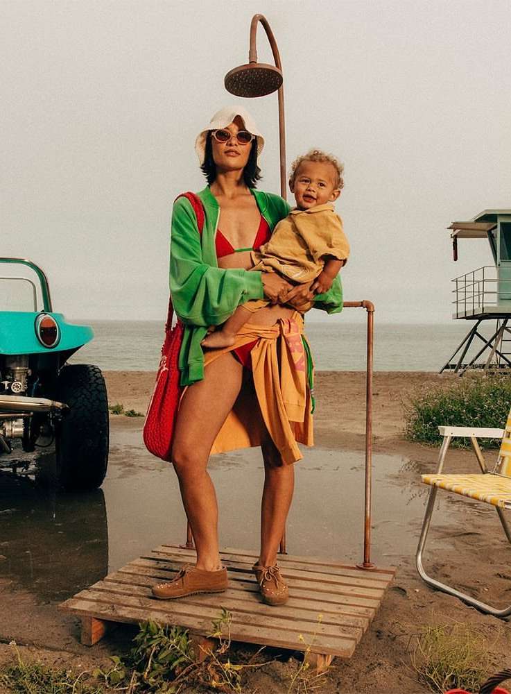 Woman holding child on a beach with a turquoise vehicle and lifeguard tower visible, under an outdoor shower.