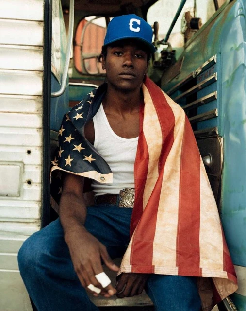 Young man draped with American flag sits casually against an aged bus, wearing a baseball cap, white tank top, jeans, and ornate belt buckle.