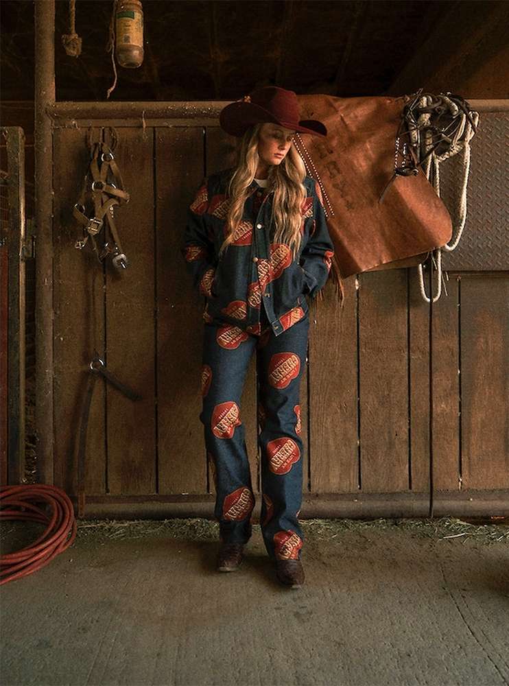 Blonde woman in a cowboy hat and patterned denim suit stands in a rustic barn with wooden walls and horse-riding equipment hanging.