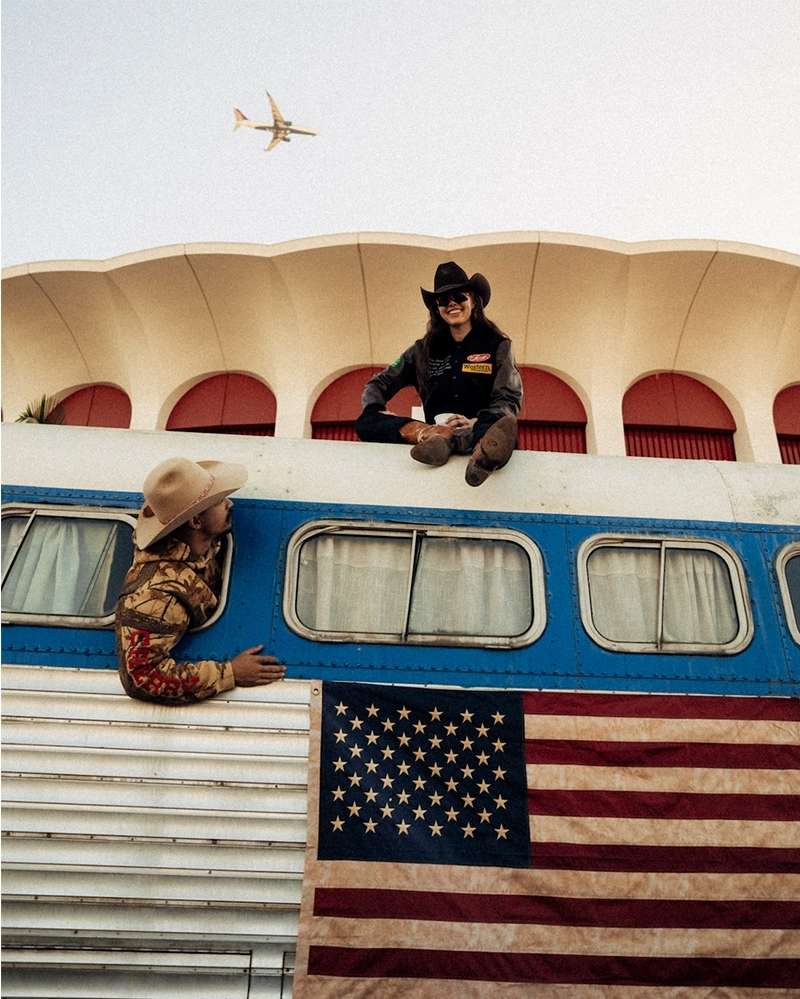 Two people in cowboy hats sit near an RV with an American flag. An airplane is flying in the background on a sunny day.