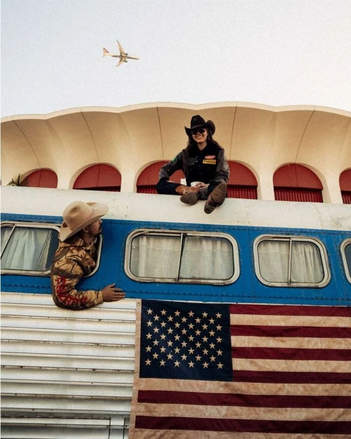 Two people in cowboy hats sit near an RV with an American flag. An airplane is flying in the background on a sunny day.