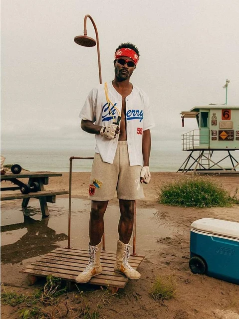 Man stands on beach with baseball jersey, shorts, boots, red bandana, sunglasses, dumbbell; shower, lifeguard tower, ocean backdrop.