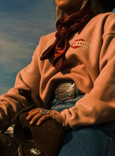 Partial view of an adult woman wearing a pink sweatshirt, jeans, ornate belt buckle, and neck scarf outdoors on a sunny day.