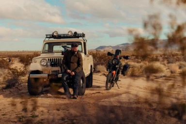 Man seated on the bumper of a vintage off-road vehicle with a parked motorcycle in a desert landscape by Nicholas Maggio