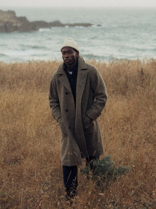 Black man wearing a brown wool coat and white beanie walking through a tall grassy field near the ocean by Nicholas Maggio