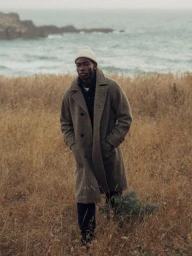 Black man wearing a brown wool coat and white beanie walking through a tall grassy field near the ocean by Nicholas Maggio