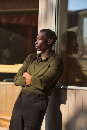 Black man in an olive green polo shirt with arms crossed leaning against a glass storefront in Los Angeles