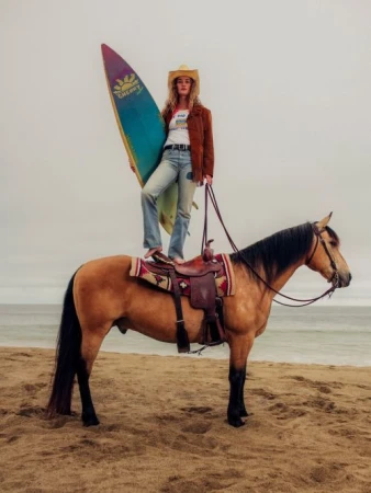 Woman wearing a cowboy hat and suede jacket standing on a horse while holding a surfboard on a sandy beach