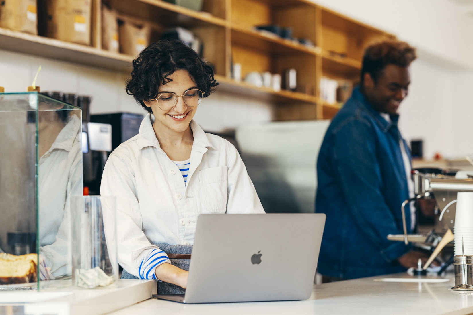 Young woman works on a laptop at a bright, modern coffee shop. Another person works at an espresso machine in the background.
