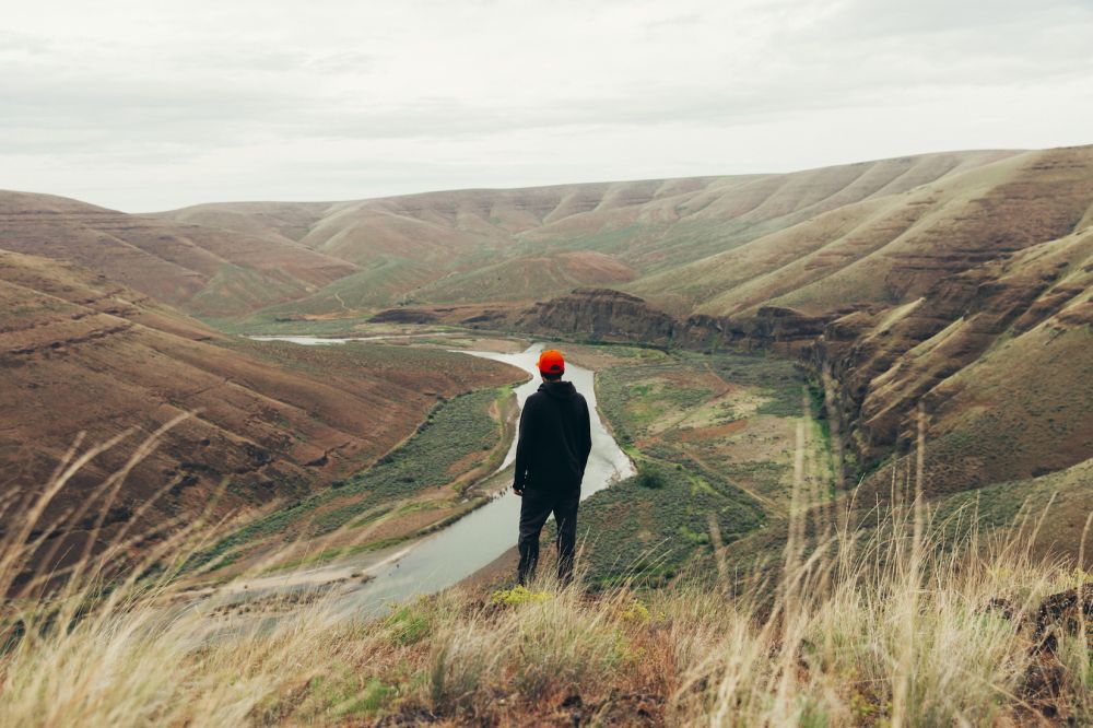 A lone hiker with a red hat gazes out over a river valley surrounded by rolling hills in a nature landscape with tall golden grass.
