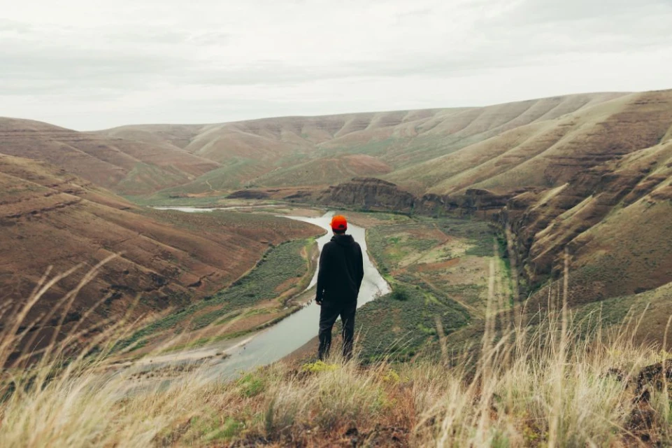 A lone hiker with a red hat gazes out over a river valley surrounded by rolling hills in a nature landscape with tall golden grass.