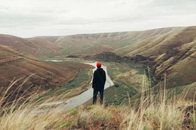 A lone hiker with a red hat gazes out over a river valley surrounded by rolling hills in a nature landscape with tall golden grass.