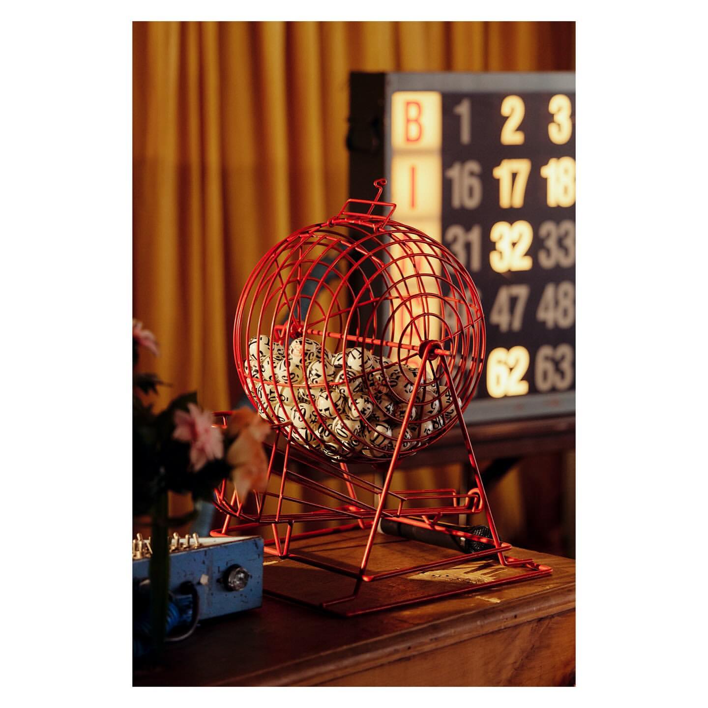 Bingo game equipment featuring a red metal bingo cage with white numbered balls, standing on a wooden table with bingo board behind it.