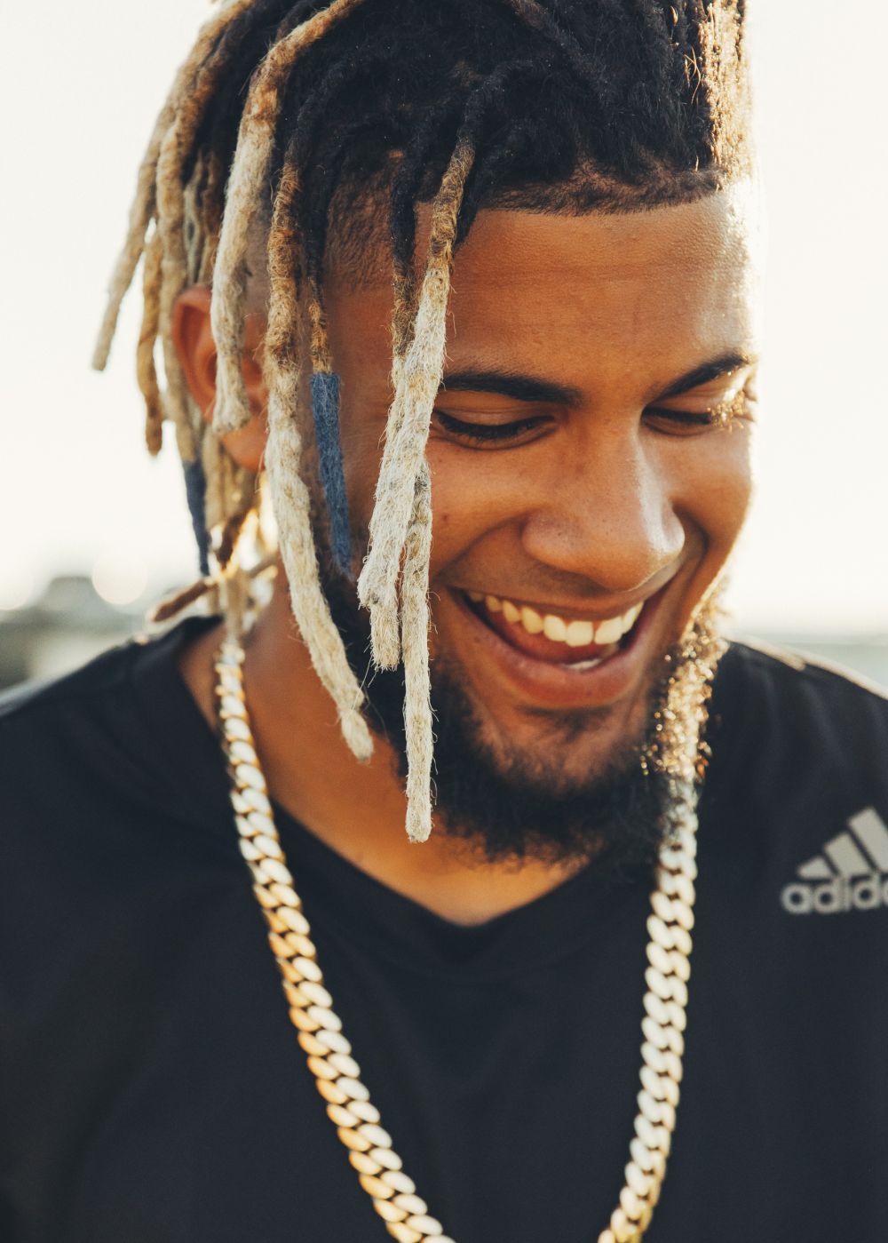 Close-up of a smiling adult male with dreadlocks and a gold chain, wearing a black t-shirt, against a bright background.