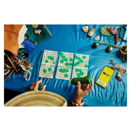 Overhead view of an adult playing bingo on a blue surface surrounded by a handbag, jewelry, and a smartphone with the Western Union app.