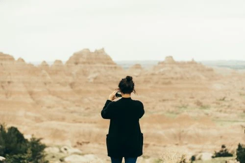 Woman taking a picture in Badlands National Park, South Dakota. The tan and brown rock formations are visible under a cloudy sky.