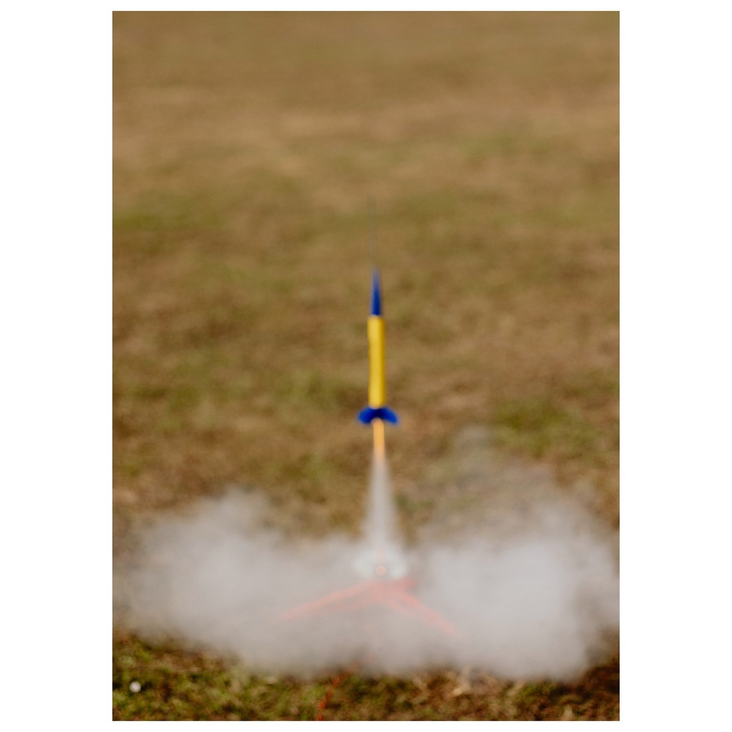 Model rocket launching outdoors with white smoke in grassy field. Yellow and blue rocket blasts off creating billows of white smoke.