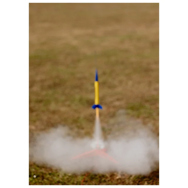 Model rocket launching outdoors with white smoke in grassy field. Yellow and blue rocket blasts off creating billows of white smoke.