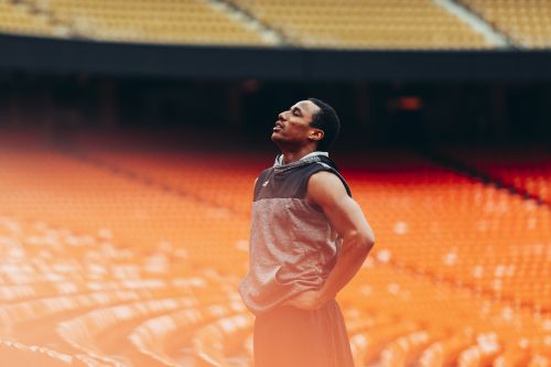 Athlete in grey sleeveless hoodie stands in an orange stadium looking up, bathed in natural light, showing a moment of calm.