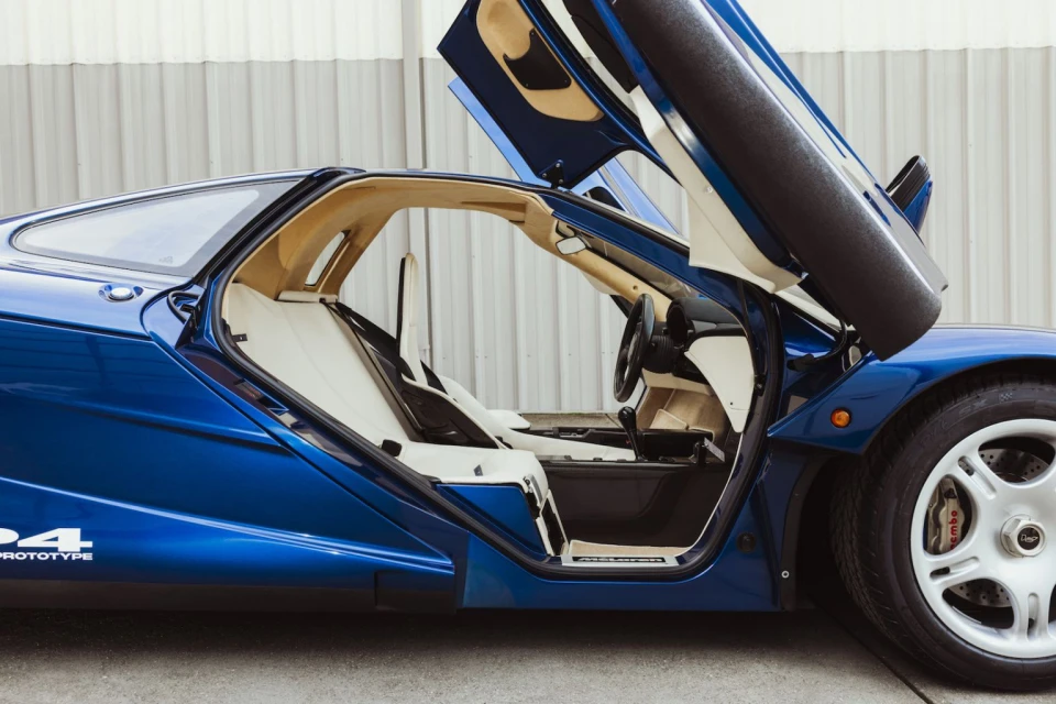 A luxurious blue McLaren F1 sports car with its gull-wing doors open, showcasing the interior and sleek design in a bright, neutral setting.