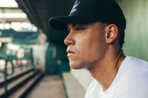 Profile portrait of a young man wearing a black baseball cap and a white t-shirt, looking forward thoughtfully in a stadium dugout.