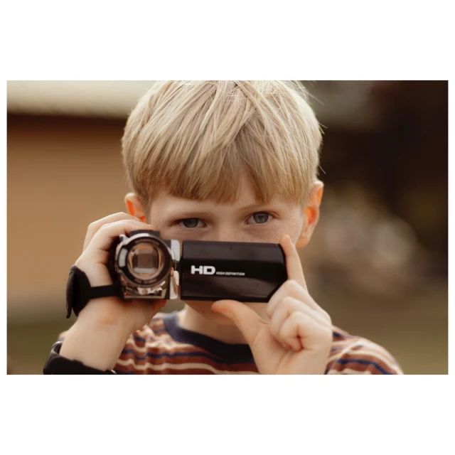 Blond-haired child films with a black handheld camcorder outdoors, looking at the camera; house visible in soft-focus background.