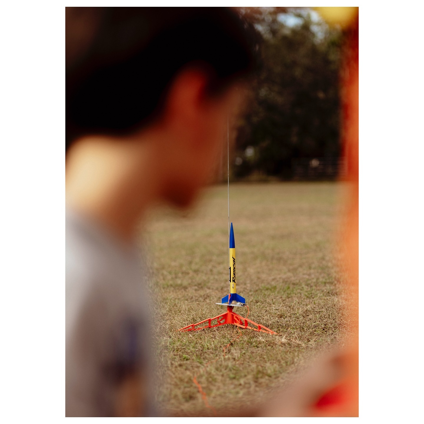 A model rocket stands on a launch pad in a grassy field, slightly blurred with an out-of-focus person in the foreground.