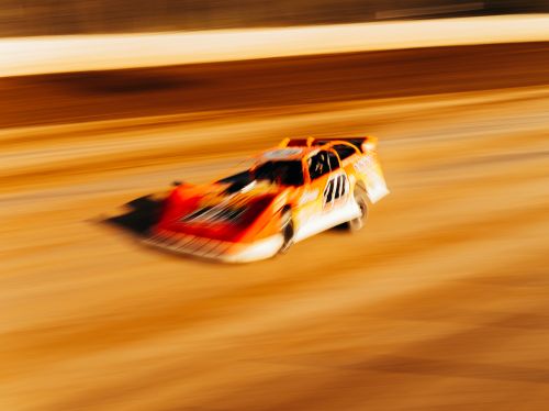 Orange and white race car speeds along a dirt track in a blurred motion, suggesting high speed and competition in motorsports.