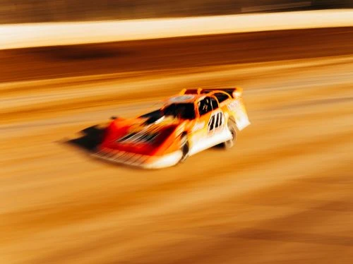 Orange and white race car speeds along a dirt track in a blurred motion, suggesting high speed and competition in motorsports.