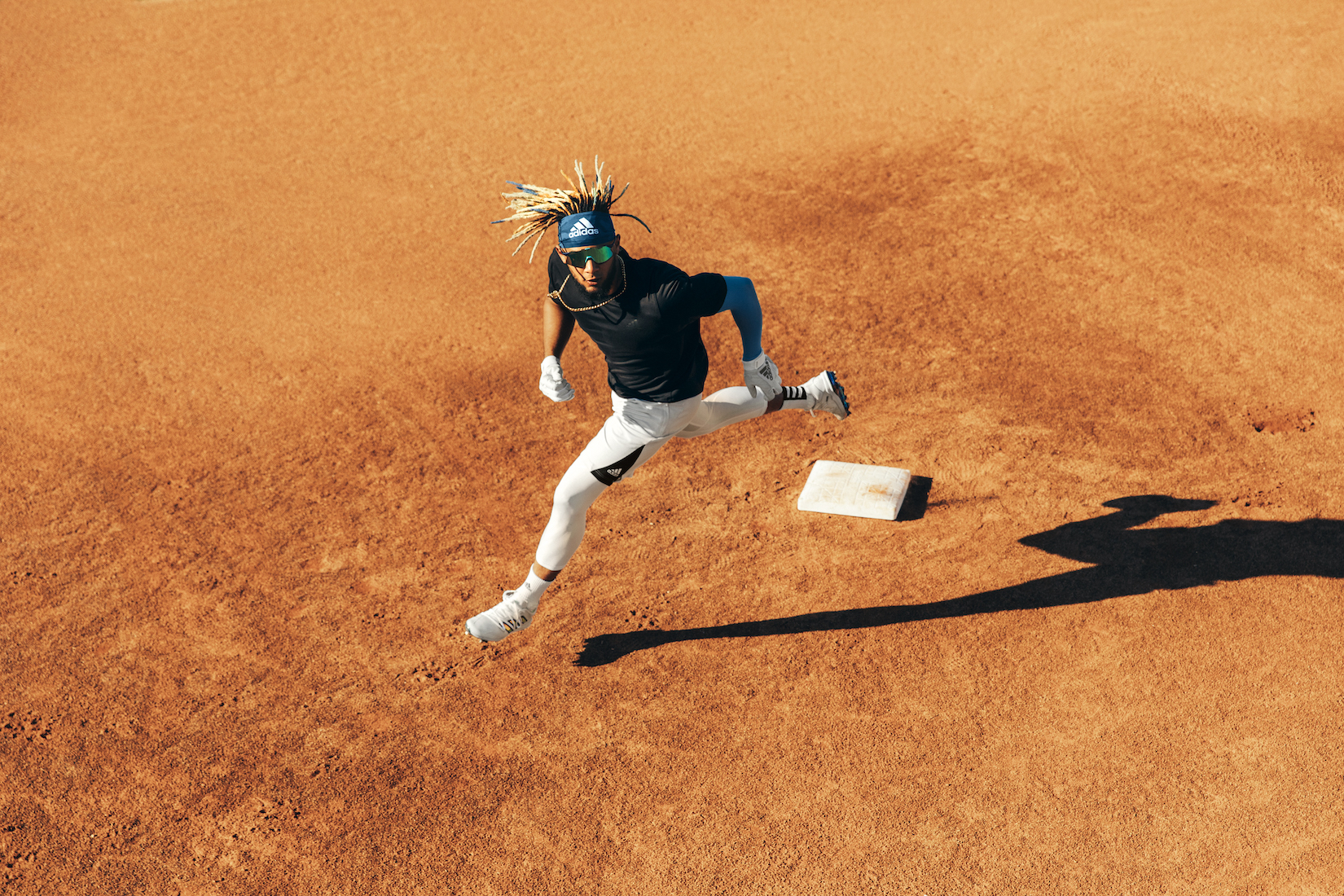 Action shot of an adult male baseball player in mid-air during a game on a dirt field, wearing Adidas apparel and sunglasses. 