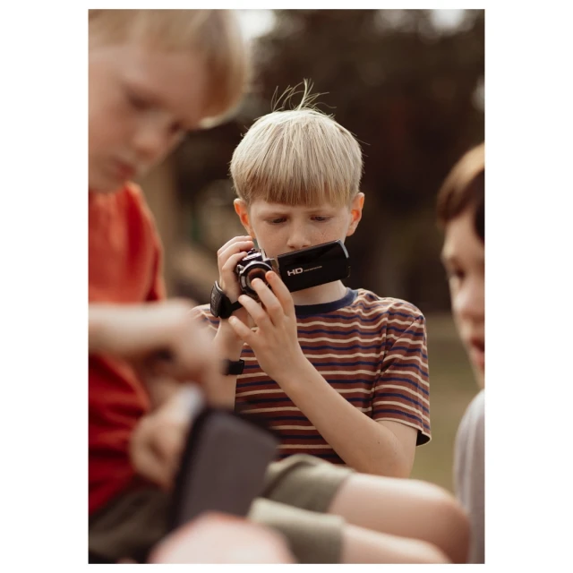 Boy films with a handheld camcorder, surrounded by other children, in an outdoor setting with natural light and shallow depth of field.