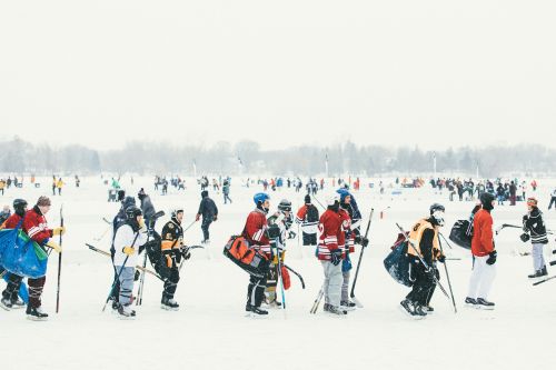 Outdoor ice hockey game with a crowd of hockey players wearing protective gear and holding hockey sticks, set on a snow covered surface.