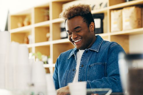 Portrait of smiling African-American man in denim jacket at cafe counter with shelves. Positive emotion, warm light, relaxed atmosphere.