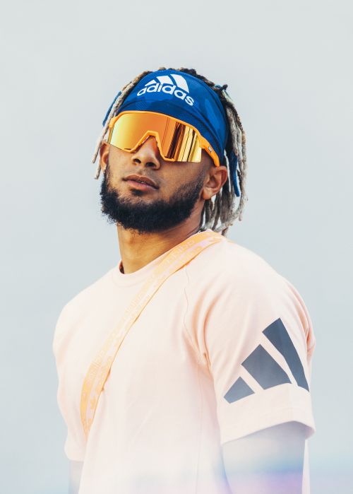 Portrait of a male adult wearing a blue Adidas headband, orange-tinted sunglasses, and a salmon-colored t-shirt, set against a light background.
